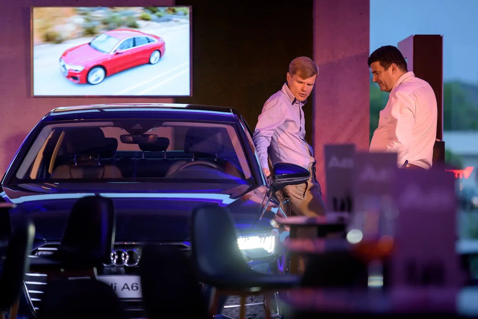 Audi A6 elölről fotózva, ahogy a lámpája világít a bemutató rendezvényen, miközben egy férfi nekitámaszkodik oldalról.