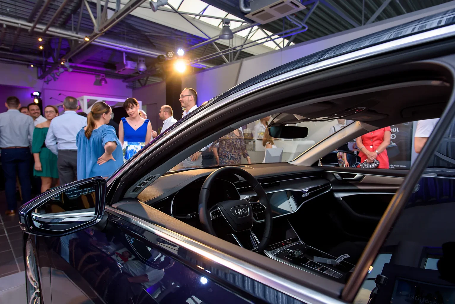Audi A6 fotó oldalról, lehúzott ablakkal a háttérben homályosan a rendezvény vendégei.